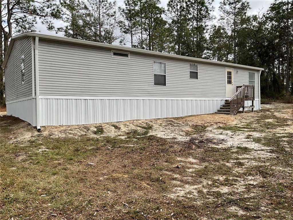 A light gray manufactured home with white skirting sits on a sandy, wooded lot — 90 NE 132ND TERRACE, WILLISTON, FL