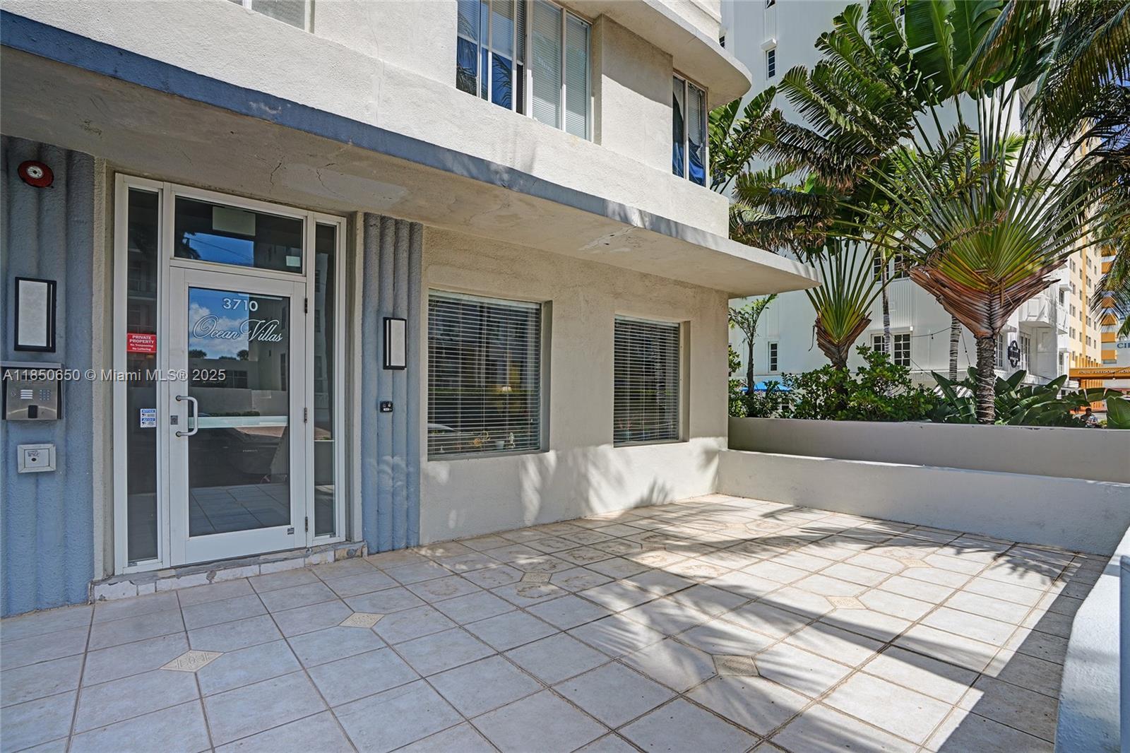 The exterior entrance of a building features a glass door, tiled patio, and palm trees — 3710 Collins Ave # 100, Miami Beach FL 33140, MIAMI BEACH, FL