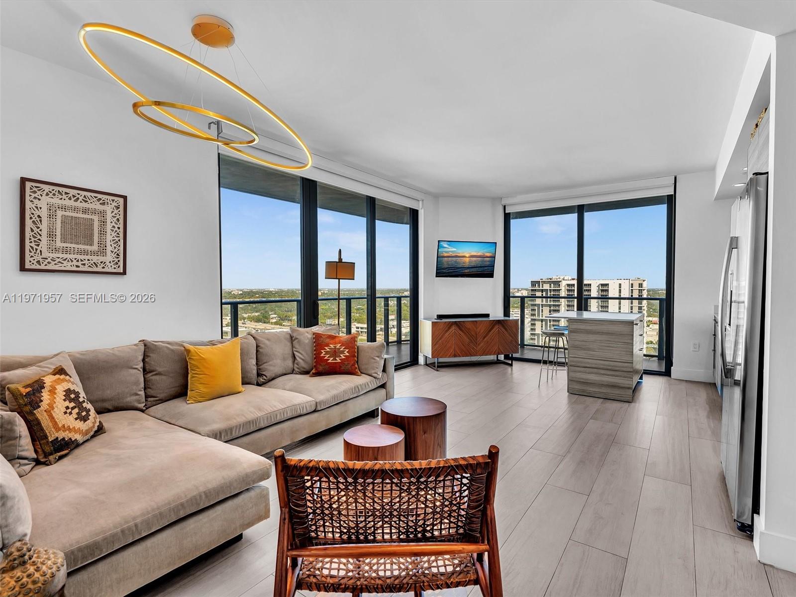 A modern living room features a sectional sofa, wood-toned flooring, and floor-to-ceiling windows with city views — 121 NE 34th St # 2016, Miami FL 33137, MIAMI, FL