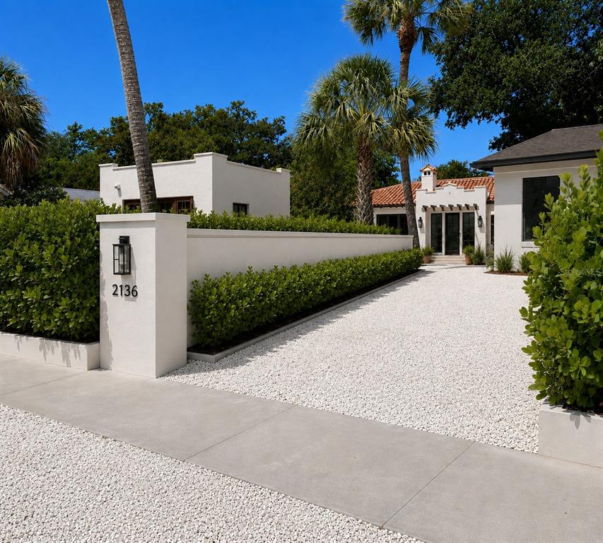 A white stucco wall and hedge line a gravel driveway leading to a Mediterranean-style home — 1041 INDIAN BEACH DRIVE, SARASOTA, FL