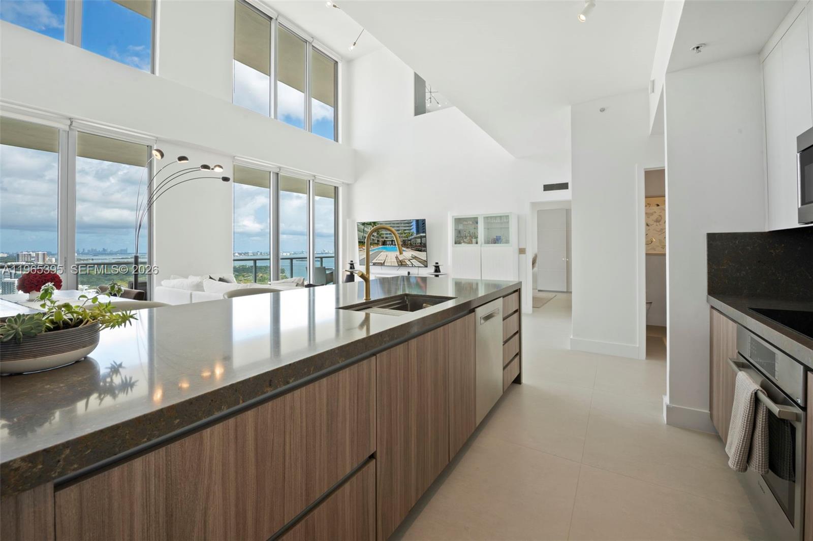 A modern kitchen features wood-grain cabinetry, dark stone countertops, and floor-to-ceiling windows with water views — 3470 E Coast Ave, Miami FL 33137, MIAMI, FL