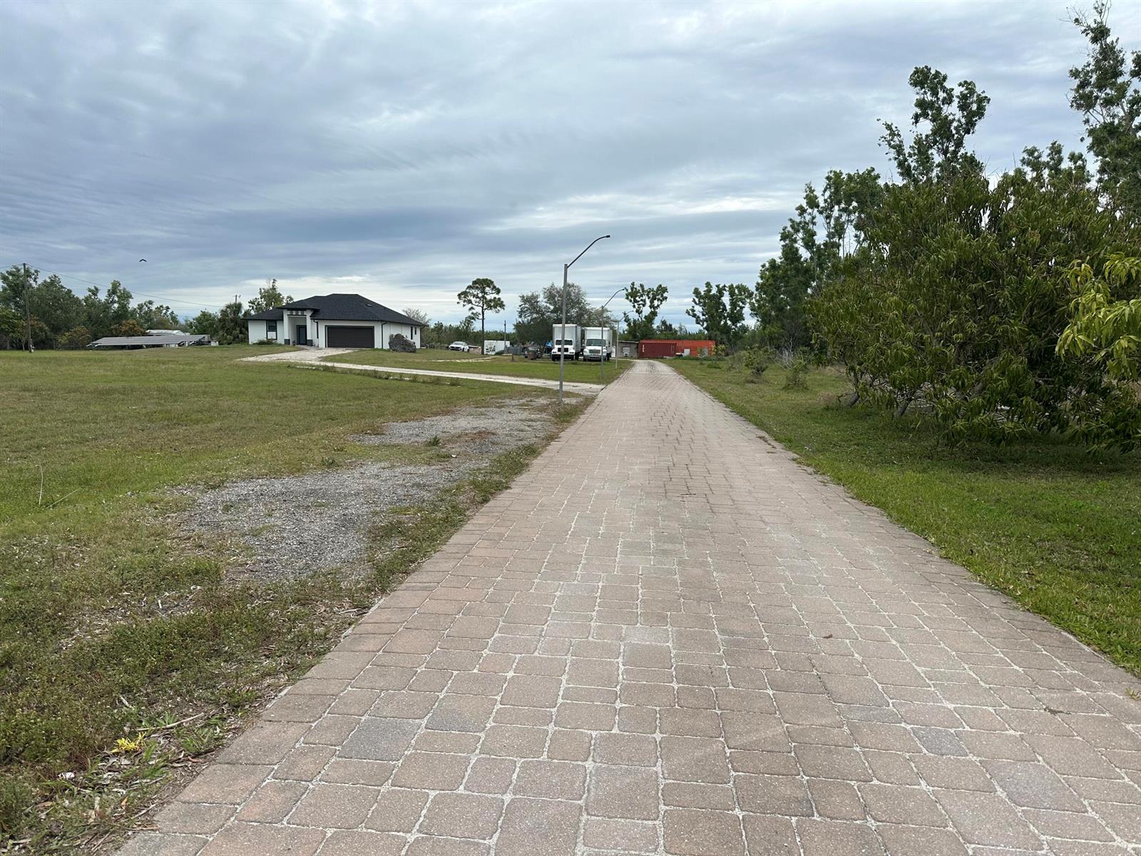 A long paver driveway leads toward a modern house under a cloudy sky — 3401 Sand Road, Cape Coral FL 33993, CAPE CORAL, FL