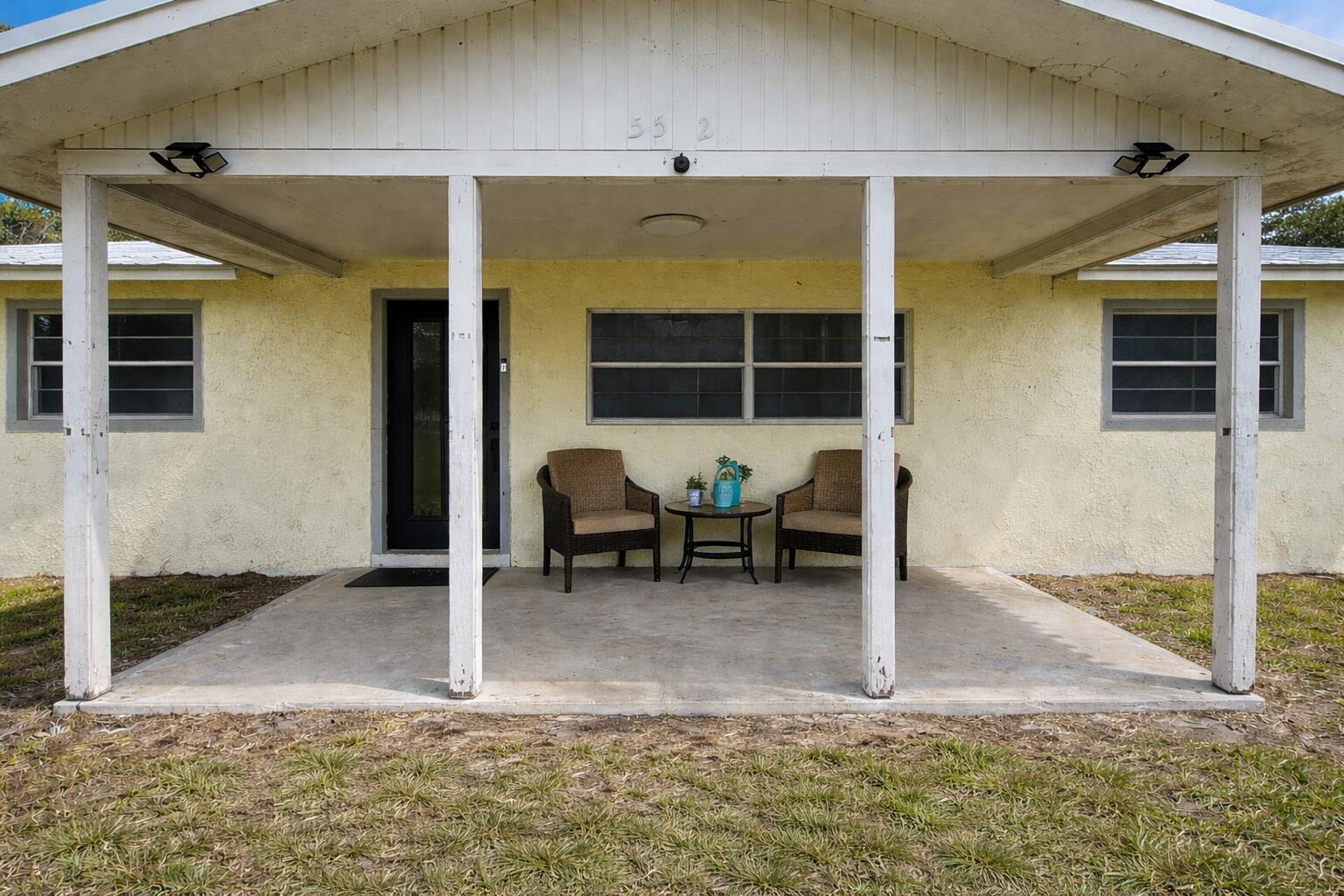 A light yellow house exterior features a covered concrete porch with two chairs and table — 5512 Buchanan Drive, Fort Pierce FL 34982, FORT PIERCE, FL
