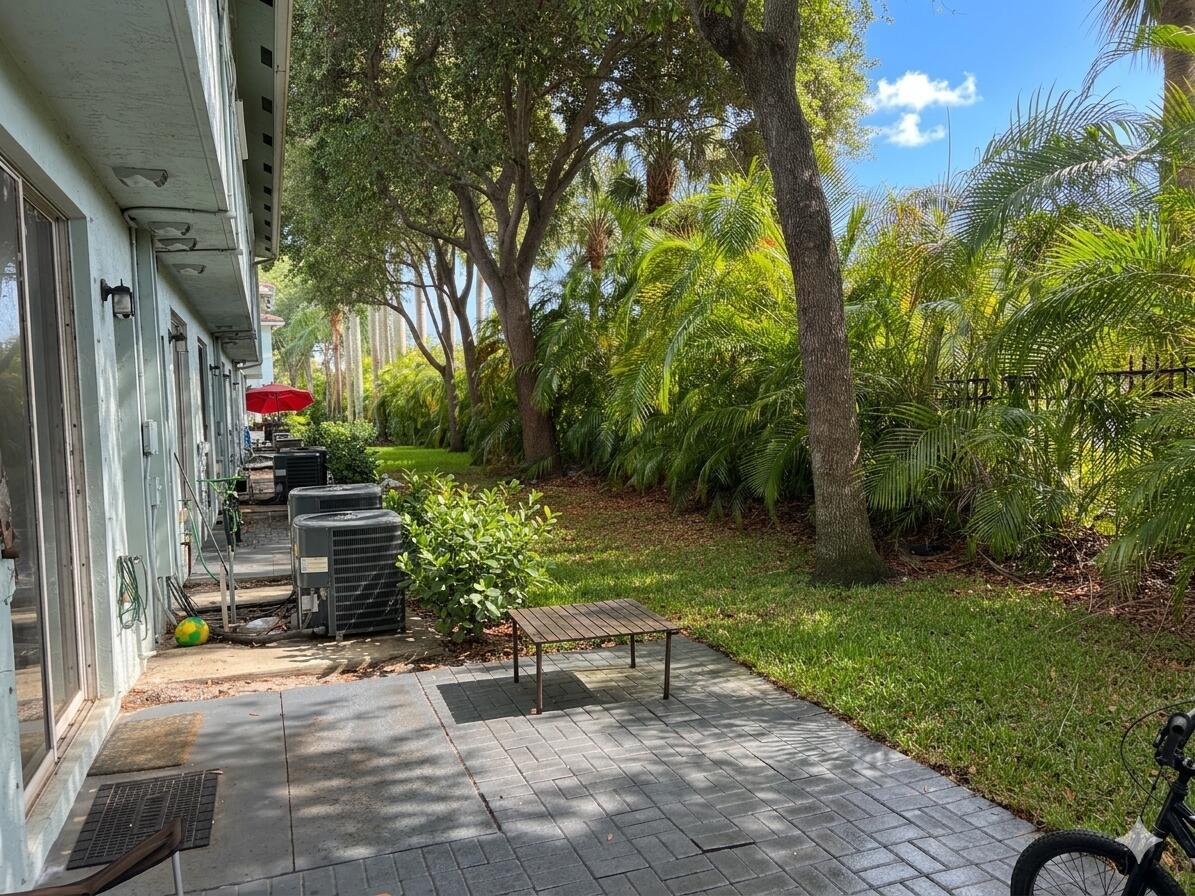 A paved patio sits beside a building exterior with air conditioning units and a grassy yard — 3731 Sonoma Drive, Riviera Beach FL 33404, RIVIERA BEACH, FL