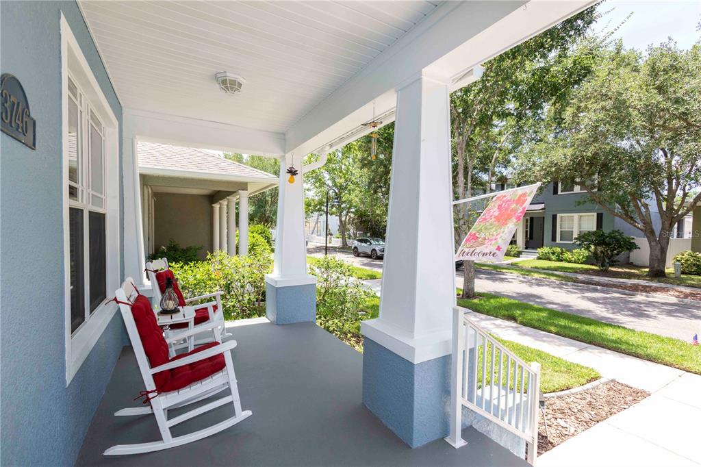 A covered front porch features white rocking chairs, blue siding, and a floral welcome flag — 3746 DOGTROT STREET, NEW PORT RICHEY, FL
