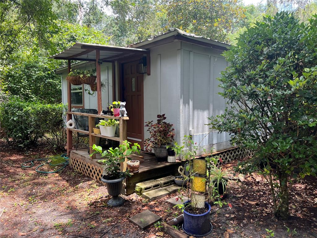 A small, light-colored shed with a wooden porch and metal roof sits in a wooded area — 6724 COUNTY ROAD 214, KEYSTONE HEIGHTS, FL