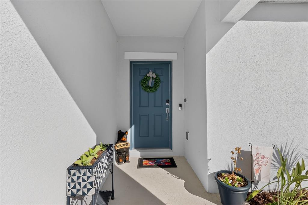 A blue front door with a wreath sits under a covered entryway with potted plants — 33692 SKY BLOSSOM CIRCLE, LEESBURG, FL