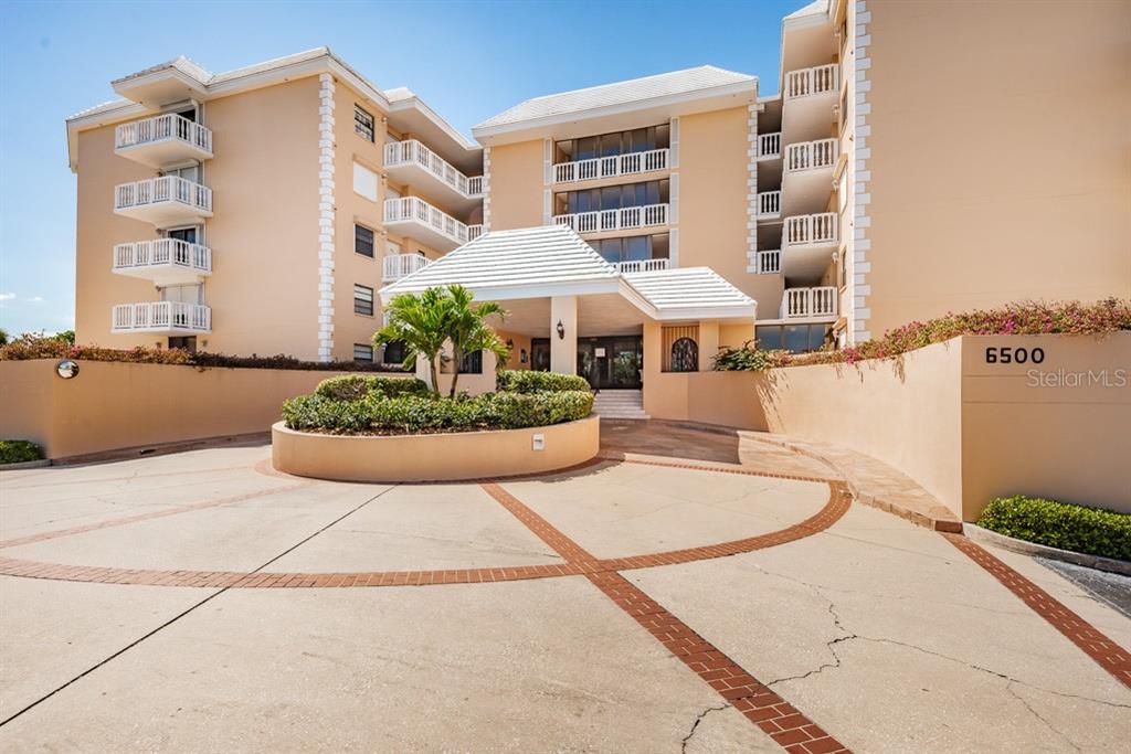 A multi-story beige apartment building features a circular driveway and a covered entrance — 6500 SUNSET WAY UNIT 505, ST PETE BEACH, FL