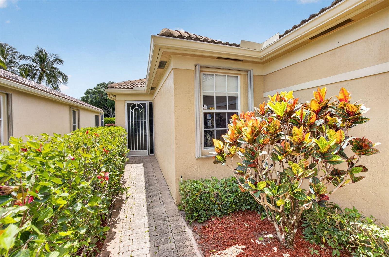 A paved walkway leads to a side entrance door beside a window and colorful landscaping — 15562 Fiorenza Circle, Delray Beach FL 33446, DELRAY BEACH, FL