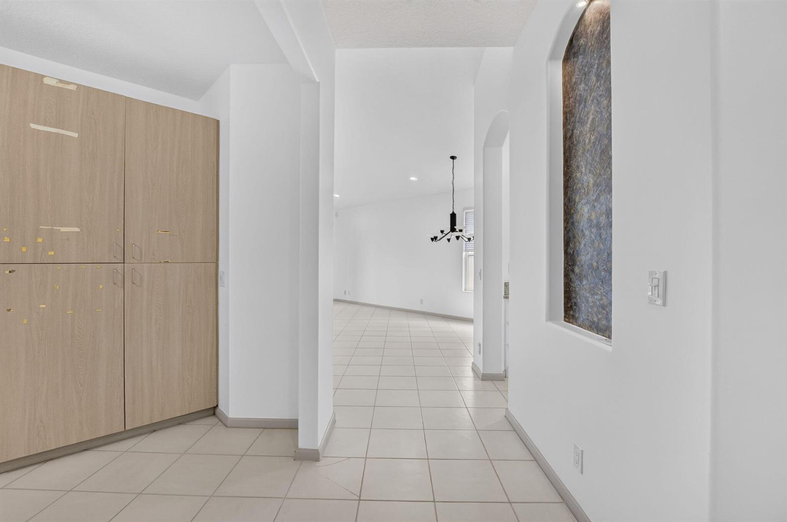 A hallway with light tile flooring leads toward a dining area with a modern chandelier — 15562 Fiorenza Circle, Delray Beach FL 33446, DELRAY BEACH, FL