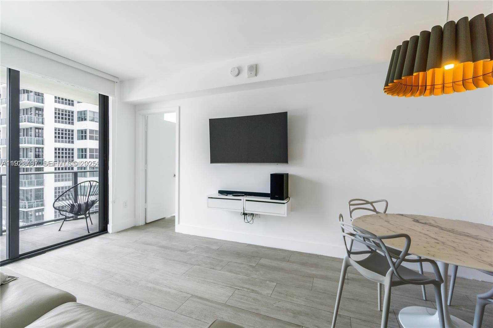 A modern living and dining area features a wall-mounted television, light wood-look flooring, and balcony — 121 NE 34th St # 1203, Miami FL 33137, MIAMI, FL