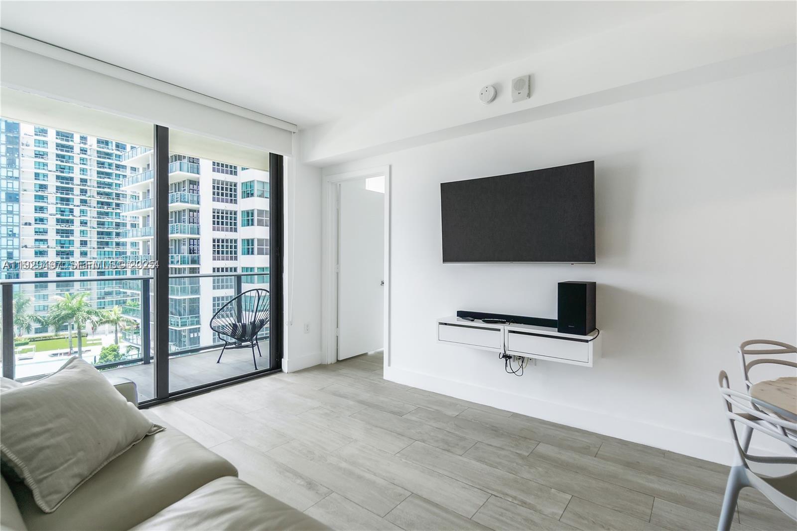 A modern living room features a wall-mounted television, floating console, and a balcony with city views — 121 NE 34th St # 1203, Miami FL 33137, MIAMI, FL