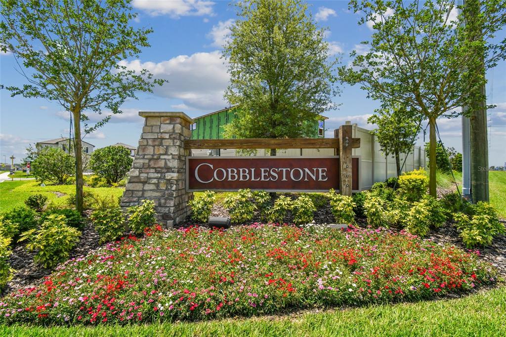 A stone monument sign for the Cobblestone community sits in a landscaped garden bed — 3211 FALL HARVEST DRIVE, ZEPHYRHILLS, FL