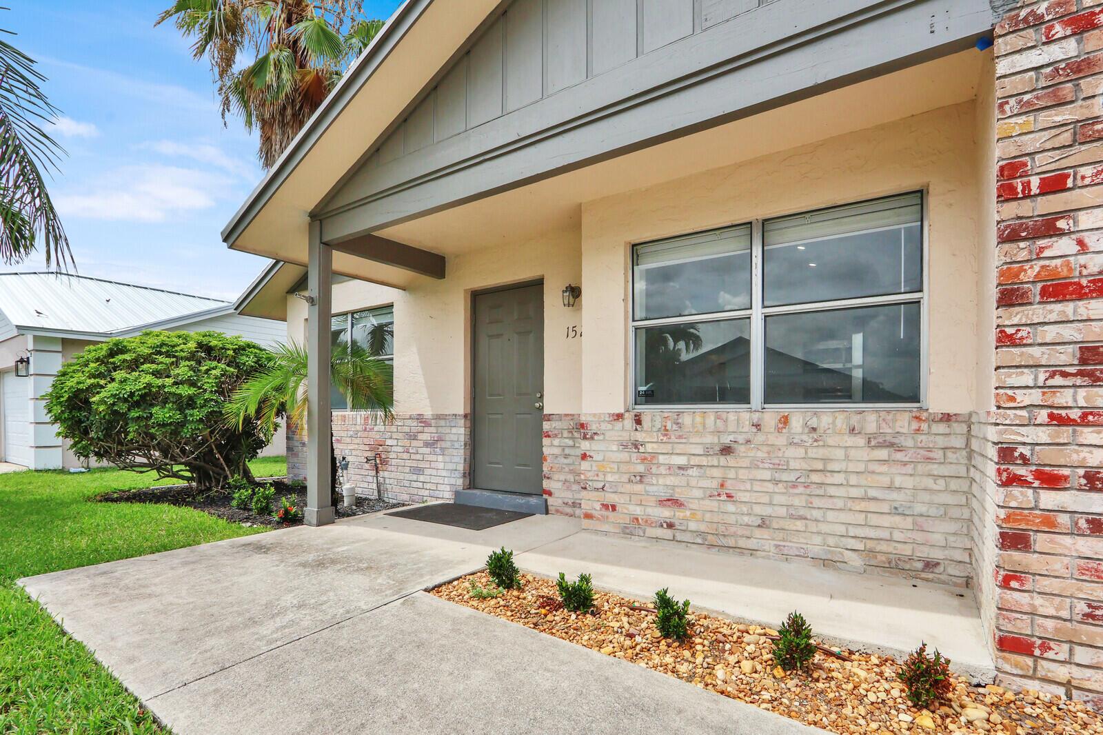 The exterior of a single-story house features a brick facade, a concrete walkway, and landscaping — 152 Wandering Trail, Jupiter FL 33458, JUPITER, FL