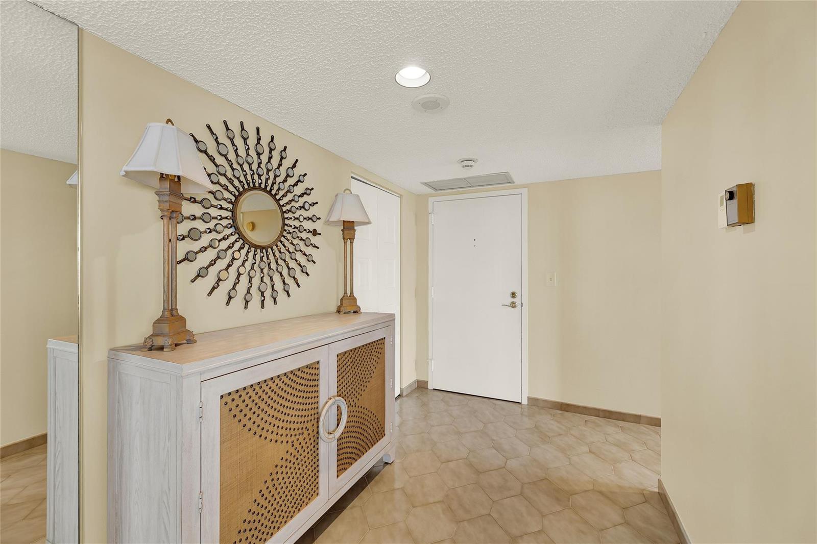 A foyer features a light-colored cabinet, a sunburst mirror, two lamps, and hexagonal floor tiles — 1010 S Ocean Boulevard # 1509, Pompano Beach FL 33062, POMPANO BEACH, FL