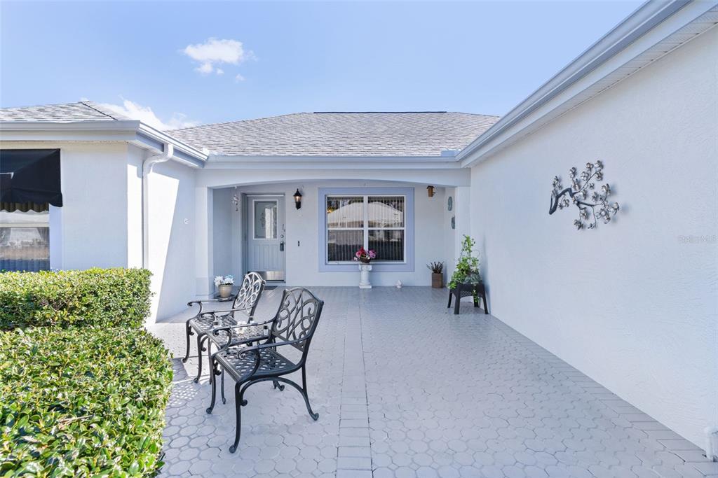 A covered patio with light-colored pavers features two metal chairs and a front entrance — 1115 SALIDO AVENUE, THE VILLAGES, FL