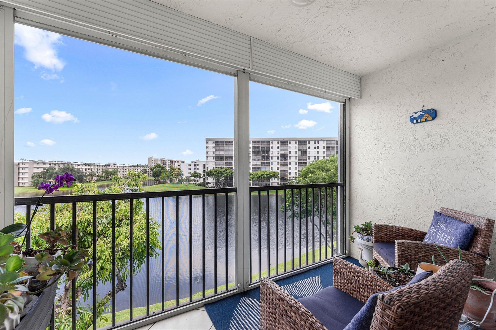 A screened-in balcony overlooks a lake and apartment buildings, featuring wicker chairs and potted plants — 2222 N Cypress Bend Drive # 403, Pompano Beach FL 33069, POMPANO BEACH, FL