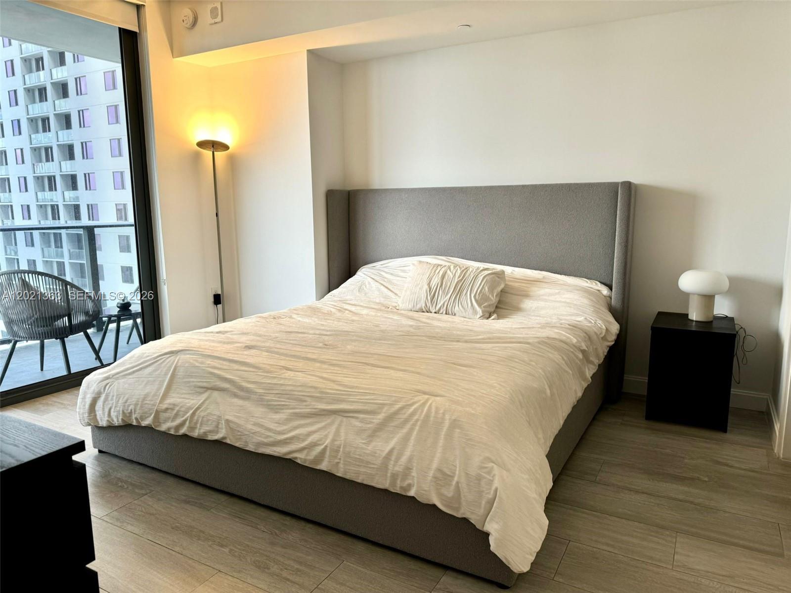A bedroom features a gray upholstered bed, a floor lamp, and a balcony with outdoor seating — 45 SW 9th St # 3107, Miami FL 33130, MIAMI, FL