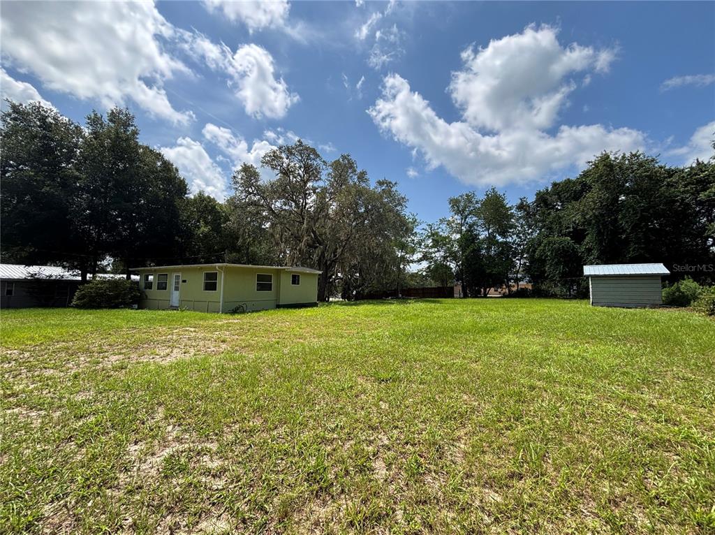 A grassy backyard features a small light green house and a detached shed under trees — 18274 SE 22ND PLACE, SILVER SPRINGS, FL