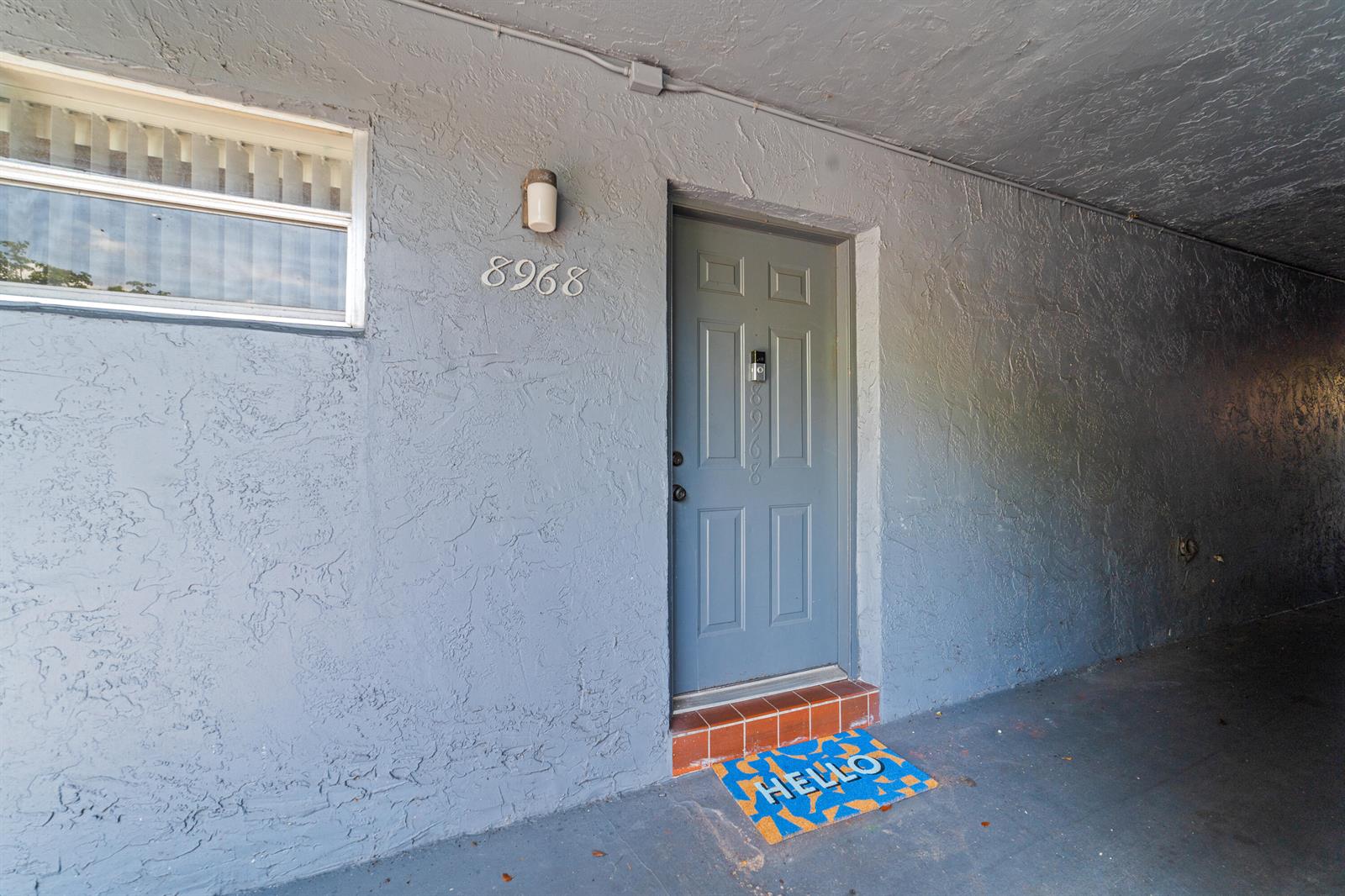 A gray exterior door with a doorbell camera sits beside a window and a doormat — 8968 W Sample Road # 101, Coral Springs FL 33065, CORAL SPRINGS, FL