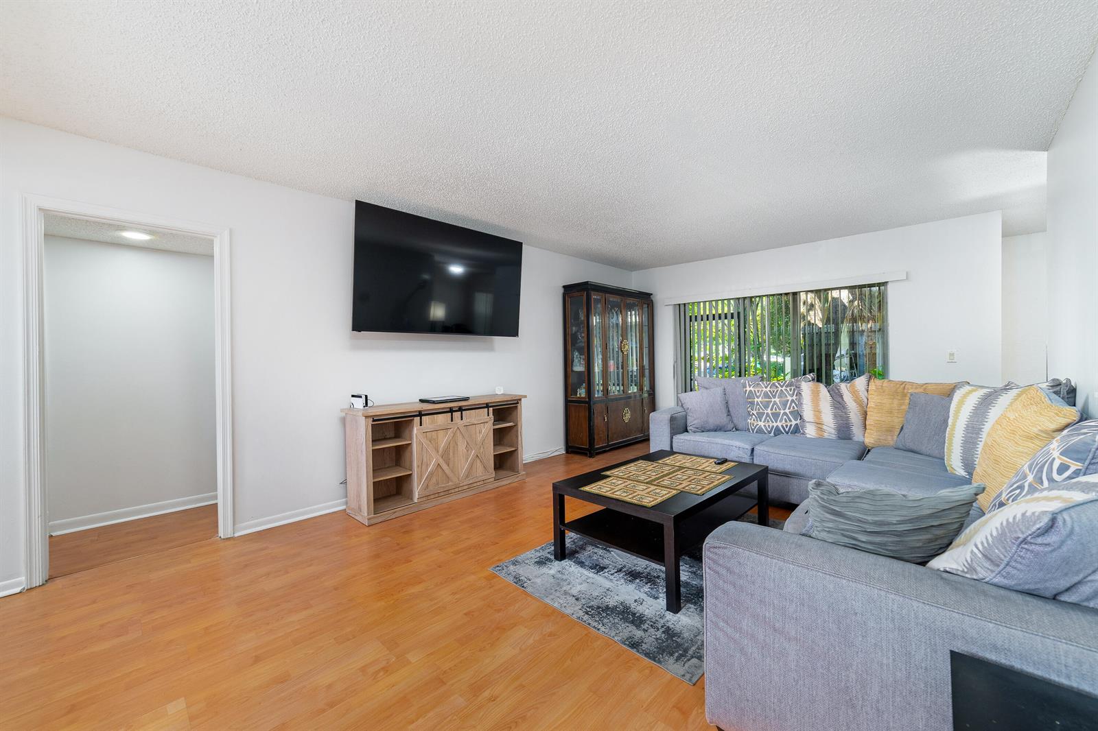 A living room features light wood flooring, a gray sectional sofa, and a wall-mounted television — 8968 W Sample Road # 101, Coral Springs FL 33065, CORAL SPRINGS, FL
