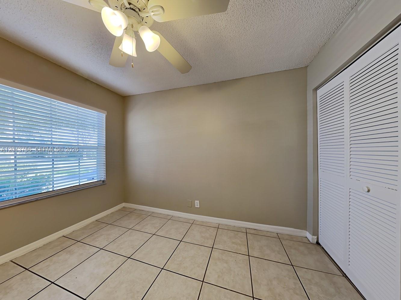 An empty room features beige walls, tiled flooring, a ceiling fan, and white louvered closet doors — MIAMI, FL
