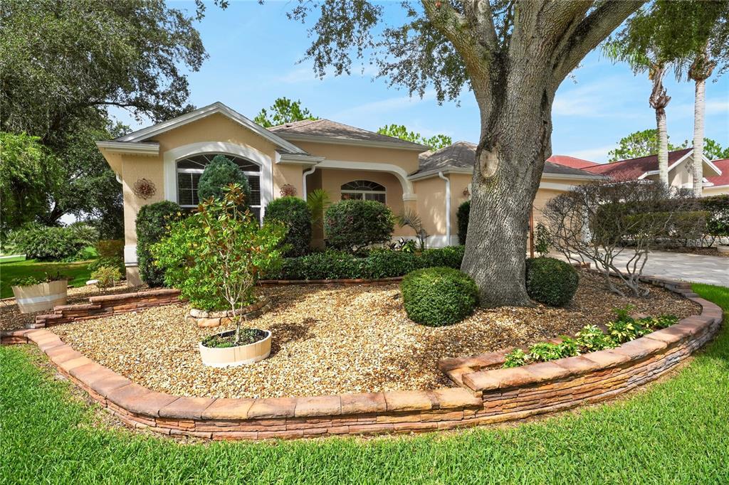 A single-story house with a tan exterior, a large oak tree, and a stone-bordered garden — 8901 SE 177TH GRASSMERE STREET, THE VILLAGES, FL