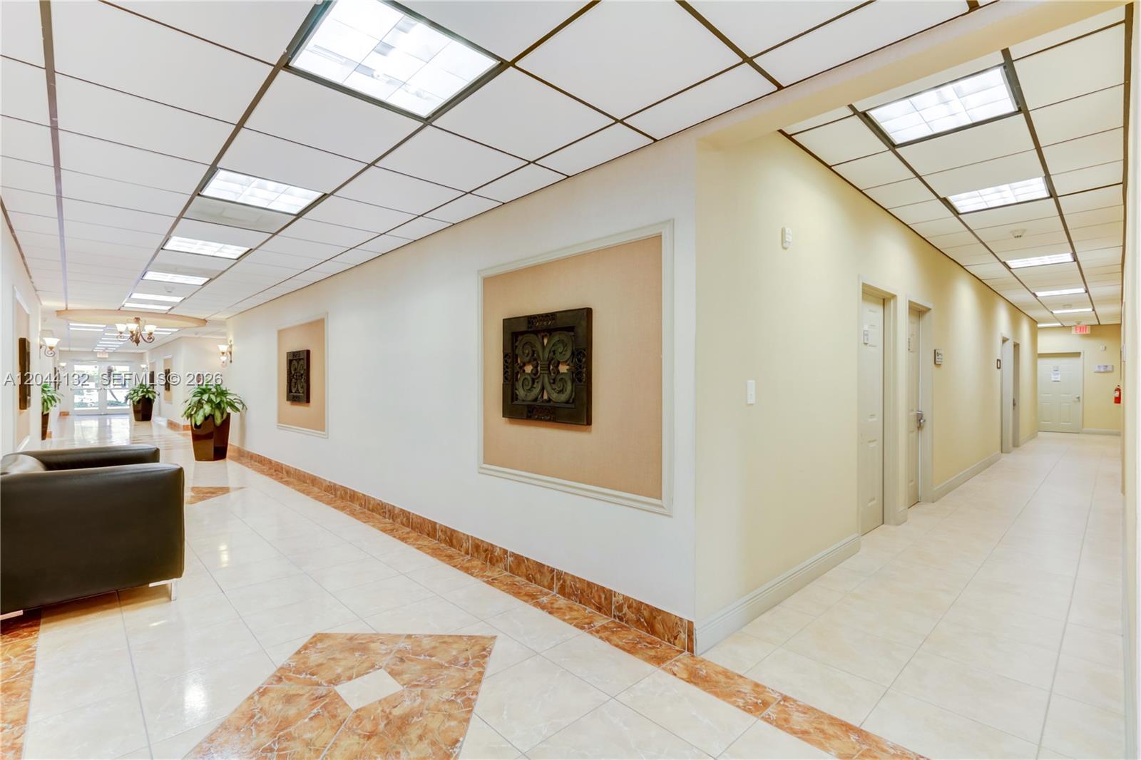 A long, brightly lit office hallway features tiled floors, drop ceilings, and decorative wall art — 12150 SW 128th Ct # 138, Miami FL 33186, MIAMI, FL
