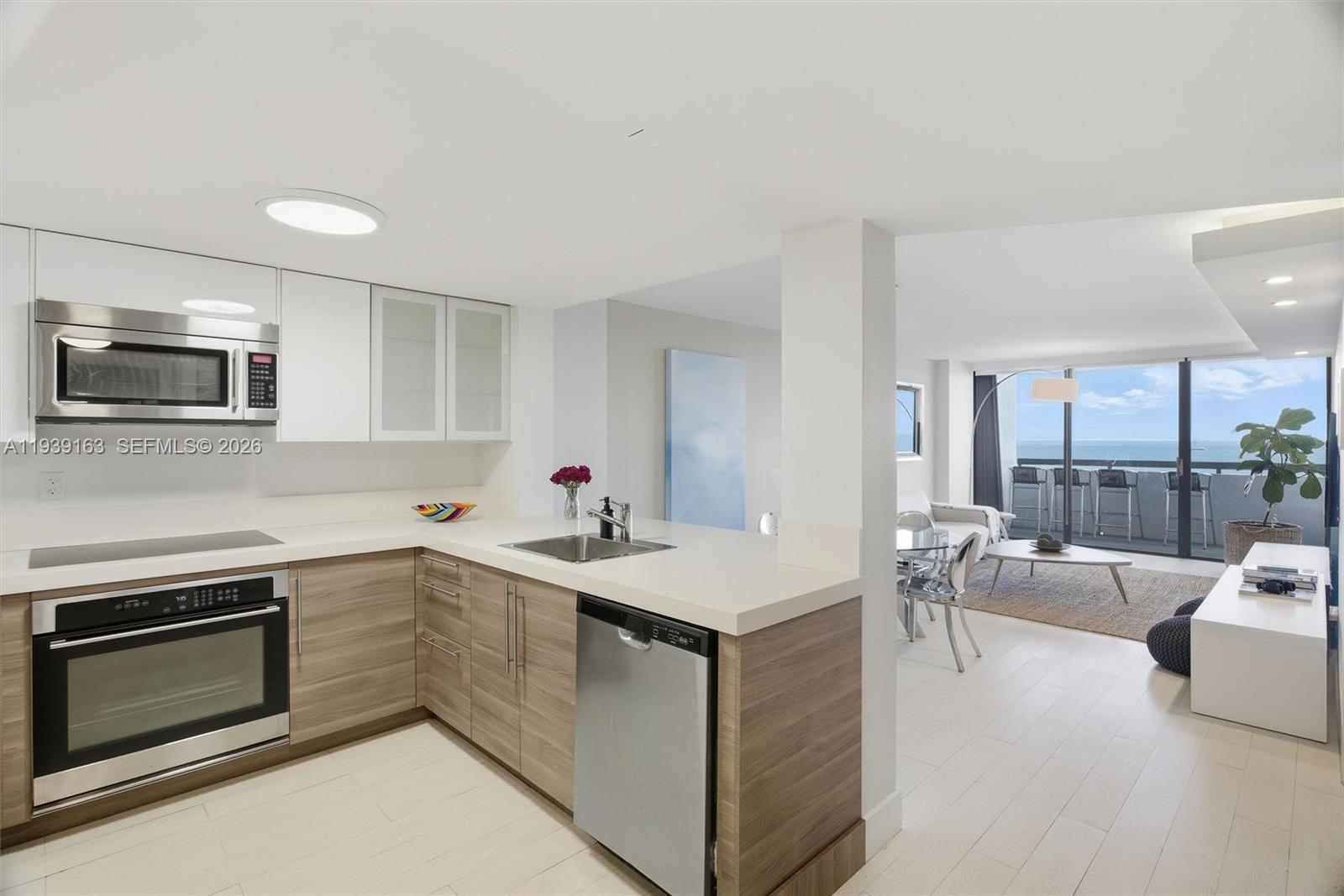 A modern kitchen with wood-tone cabinets and white countertops opens to a bright living area — 2555 Collins Ave # 2105, Miami Beach FL 33140, MIAMI BEACH, FL