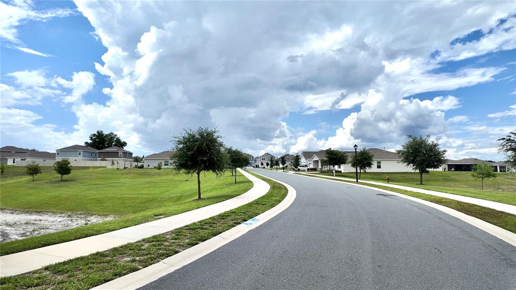 A paved road curves through a residential neighborhood with houses and green lawns under clouds — 5620 WINDSONG OAK DRIVE, LEESBURG, FL