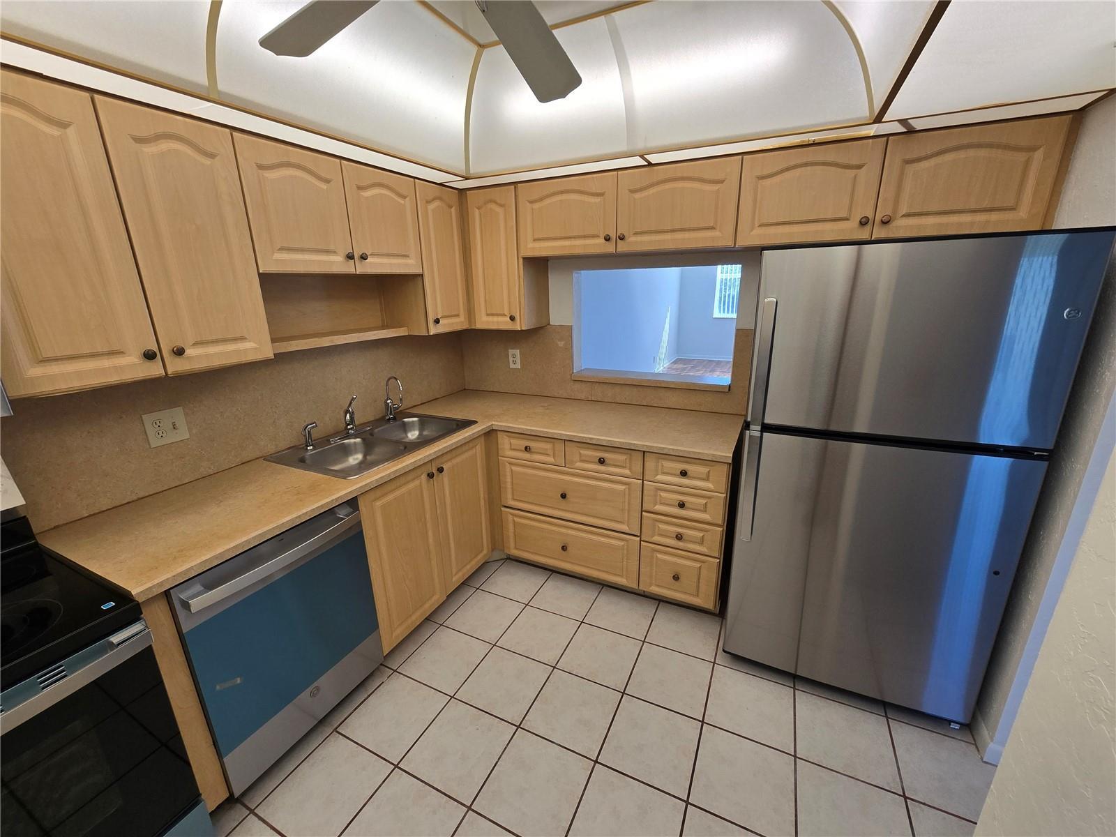 A kitchen features light wood cabinets, stainless steel appliances, beige countertops, and a pass-through window — 6890 Royal Palm Boulevard # 211h, Margate FL 33063, MARGATE, FL