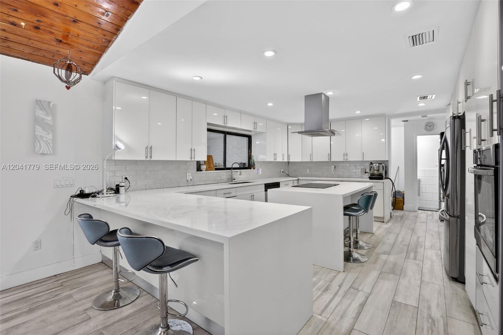 A modern kitchen features white cabinetry, a large waterfall island, and light wood-look flooring — DAVIE, FL