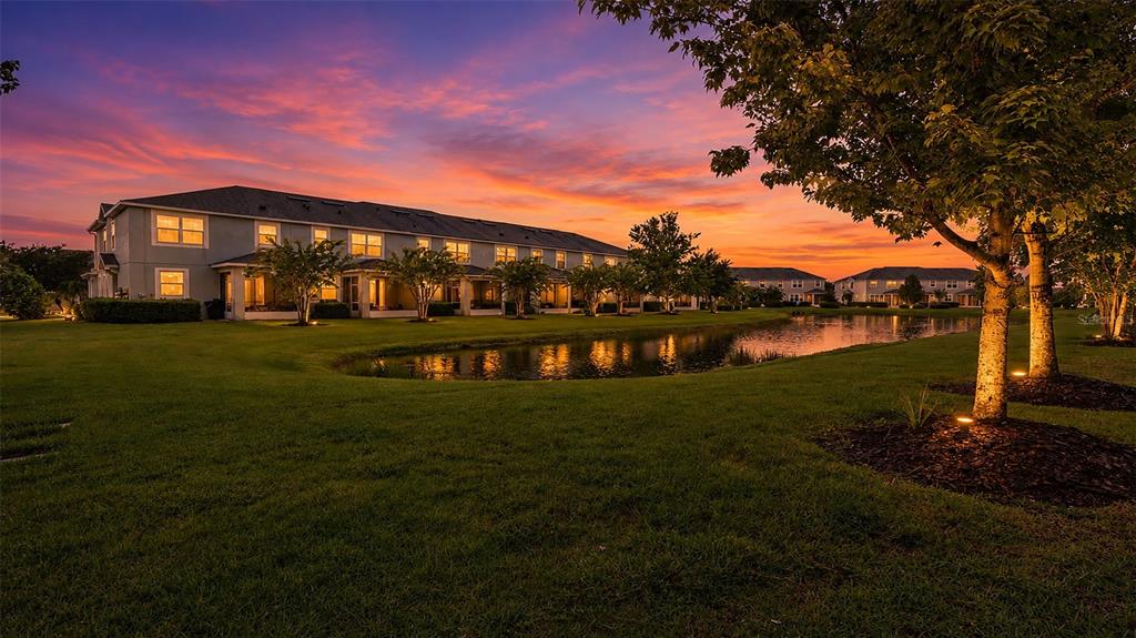 A multi-unit residential building sits beside a pond at sunset with trees in the foreground — 12356 TRAILHEAD DRIVE, BRADENTON, FL