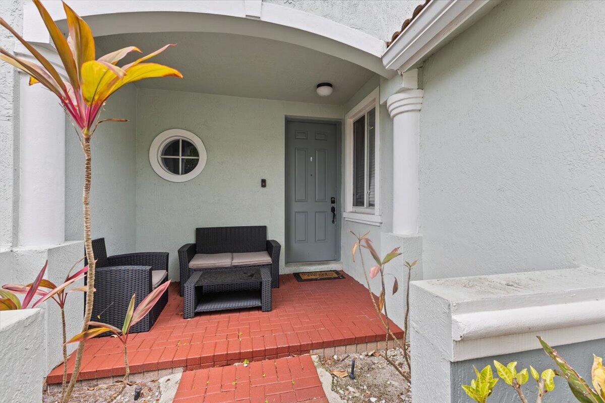 A covered porch features red brick flooring, a gray front door, and wicker patio furniture — 2227 Oakmont Drive, Riviera Beach FL 33404, RIVIERA BEACH, FL