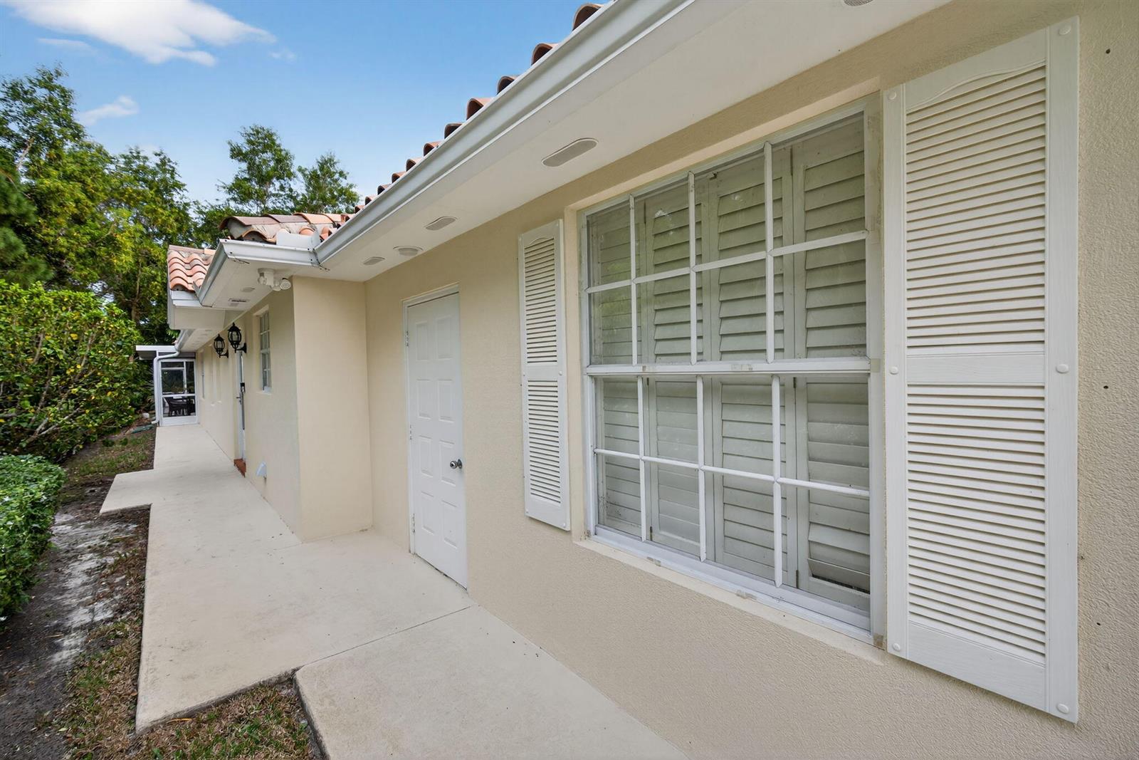 A beige exterior wall features a white door, a window with shutters, and a walkway — 8381 SE Double Tree Drive, Hobe Sound FL 33455, HOBE SOUND, FL