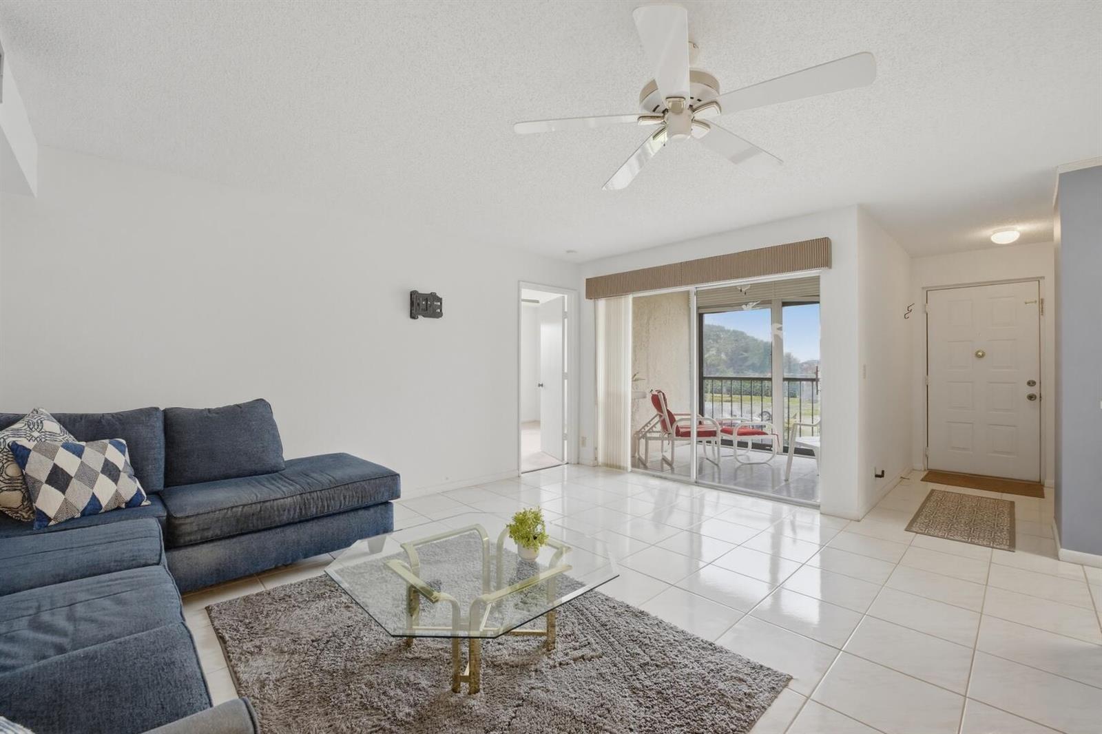 A living room features white tile floors, a blue sectional sofa, and a sliding glass door — 14898 Wedgefield Drive # 208, Delray Beach FL 33446, DELRAY BEACH, FL