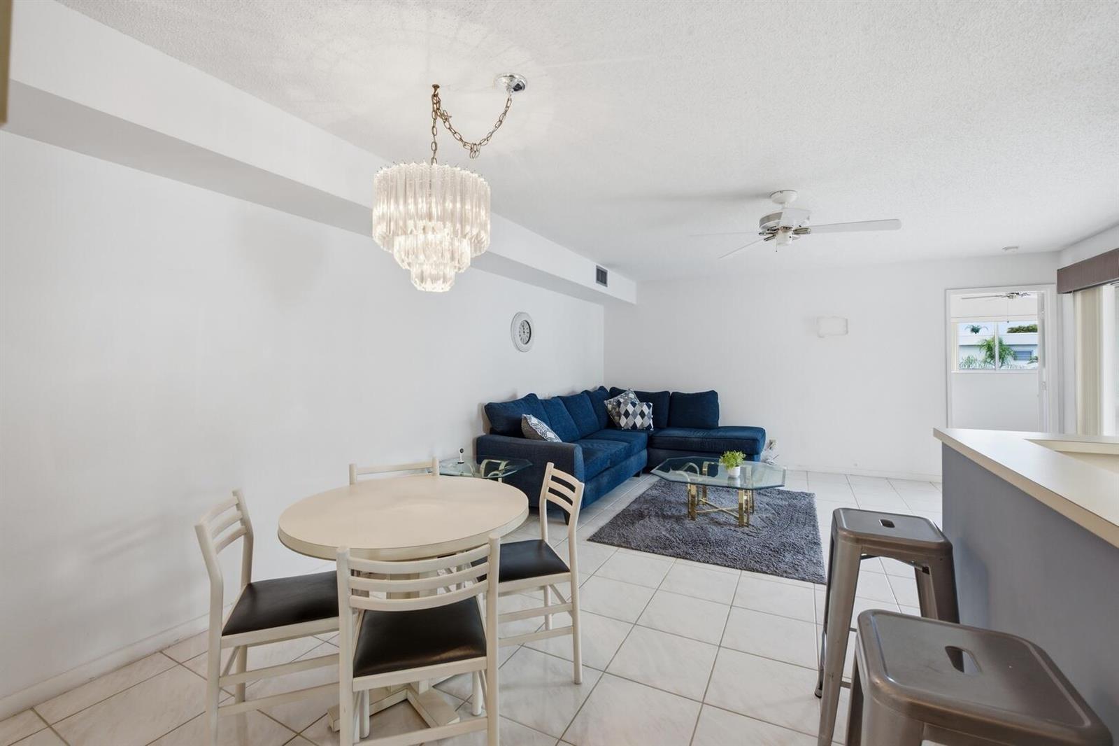 An open-concept living and dining area features white tile floors, a blue sectional, and chandelier — 14898 Wedgefield Drive # 208, Delray Beach FL 33446, DELRAY BEACH, FL