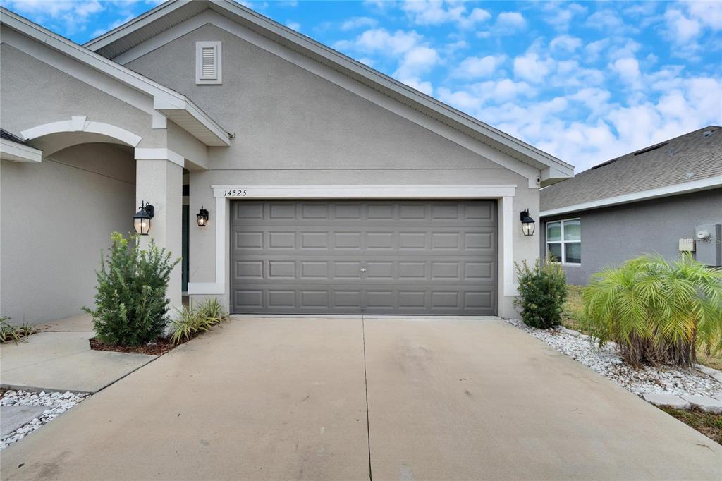 A gray stucco house features a two-car garage, a concrete driveway, and exterior lighting — 14525 SCOTTBURGH GLEN DRIVE, WIMAUMA, FL