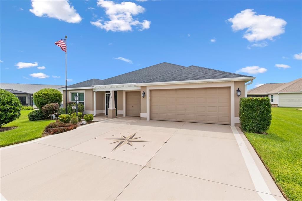 A single-story house with a beige exterior, a two-car garage, and a compass-patterned concrete driveway — 5996 DELPHINA LOOP, THE VILLAGES, FL