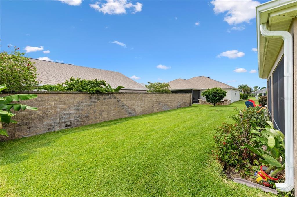 A grassy backyard features a long concrete block wall and a screened-in patio area — 5996 DELPHINA LOOP, THE VILLAGES, FL