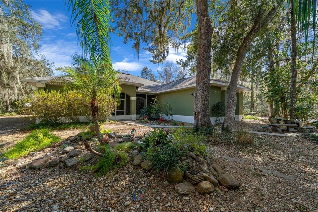 A single-story green house with a screened entryway sits in a wooded, gravel-landscaped yard — 16731 SE 181ST TERRACE, WEIRSDALE, FL