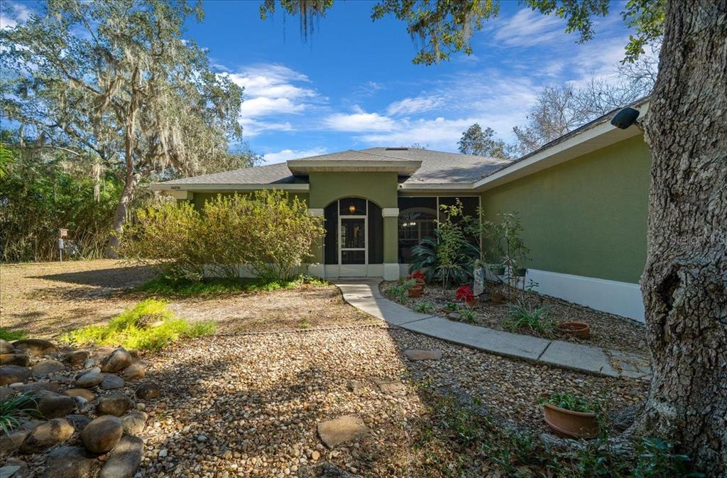 A single-story house with green stucco walls, a screened entryway, and a gravel front yard — 16731 SE 181ST TERRACE, WEIRSDALE, FL