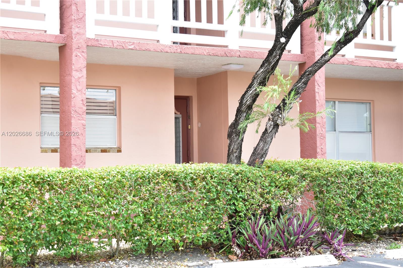 A pink building exterior features a covered entryway, windows, and a manicured green hedge — 9461 Sunrise Lakes Blvd # 103, Sunrise FL 33322, SUNRISE, FL