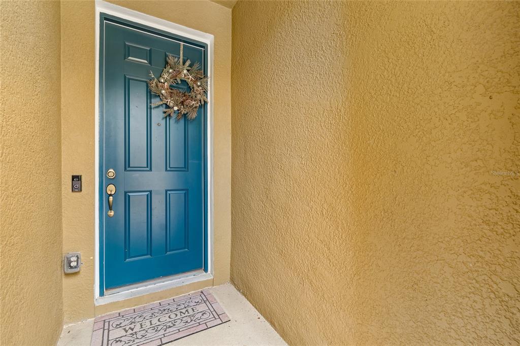 A teal front door with a wreath and a welcome mat sits in a stucco entryway — 17852 ALTHEA BLUE PLACE, LUTZ, FL