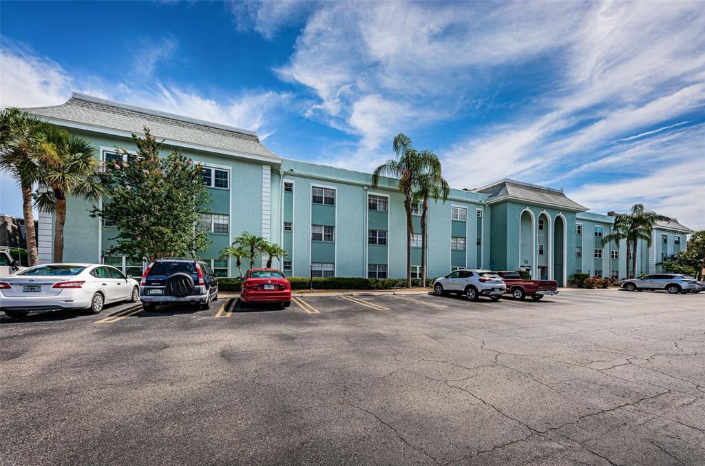 A multi-story teal apartment building stands behind a parking lot with several parked cars — 1706 BELLEAIR FOREST DRIVE UNIT 322, BELLEAIR, FL