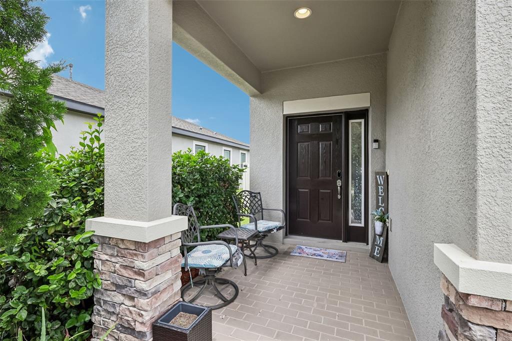 A covered front porch features a dark wood door, two metal chairs, and stone-accented columns — 8210 MALLOW MIRROR LANE, LAND O LAKES, FL