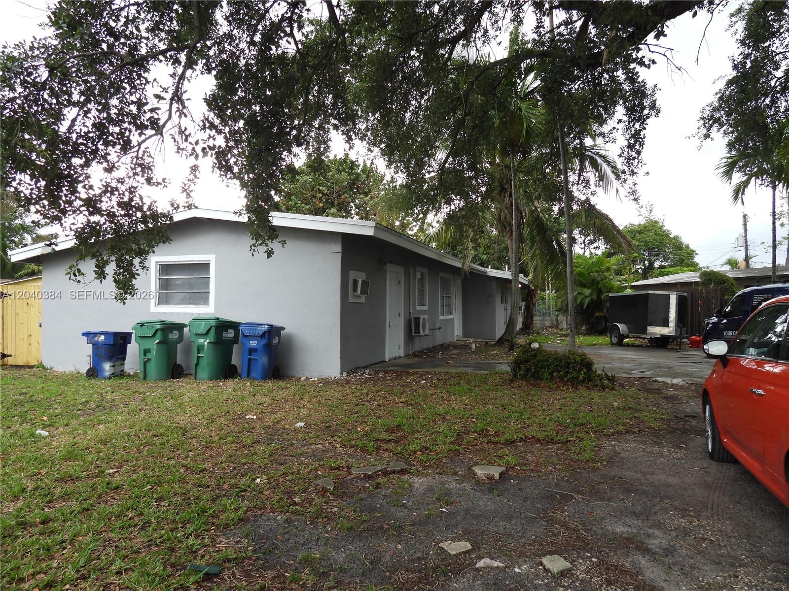 A gray, single-story house exterior features four trash bins and a parked red car — MIAMI, FL