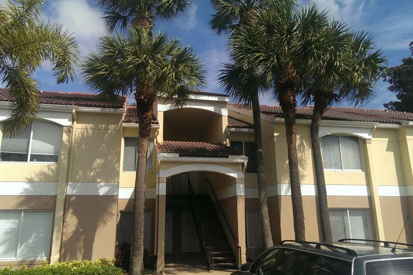 A two-story apartment building exterior features beige stucco walls, palm trees, and a central staircase — 815 W Boynton Beach Boulevard # 204, Boynton Beach FL 33426, BOYNTON BEACH, FL