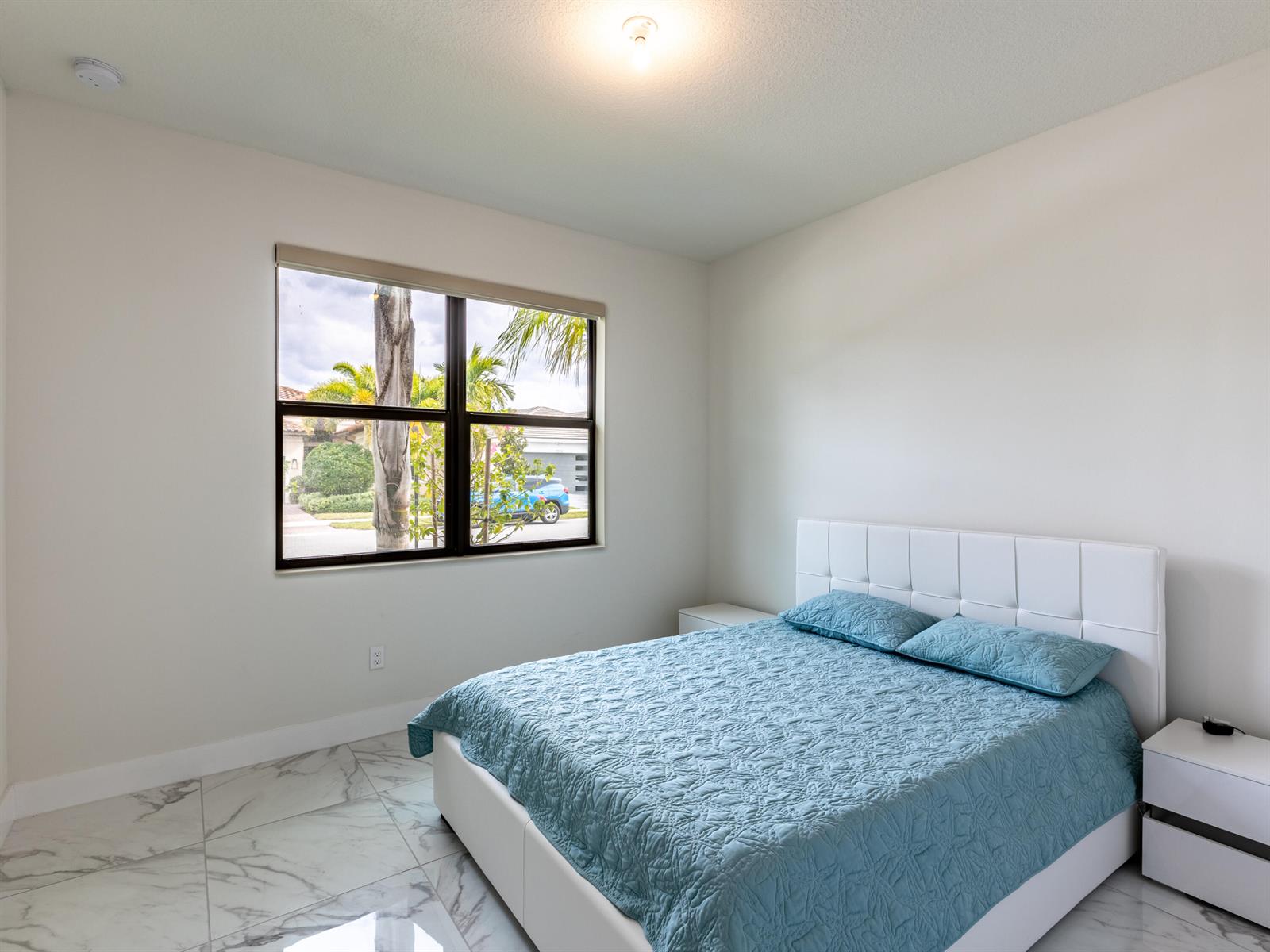 A bedroom features a white bed with a blue quilt, marble floors, and a window — 7191 Montereal Path, Lake Worth FL 33463, LAKE WORTH, FL