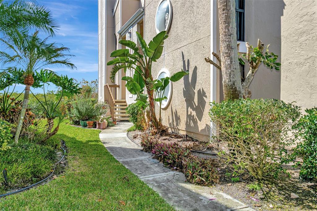 A concrete walkway leads past a tan building exterior with round windows and tropical landscaping — 8734 54TH AVENUE W UNIT 8734, BRADENTON, FL
